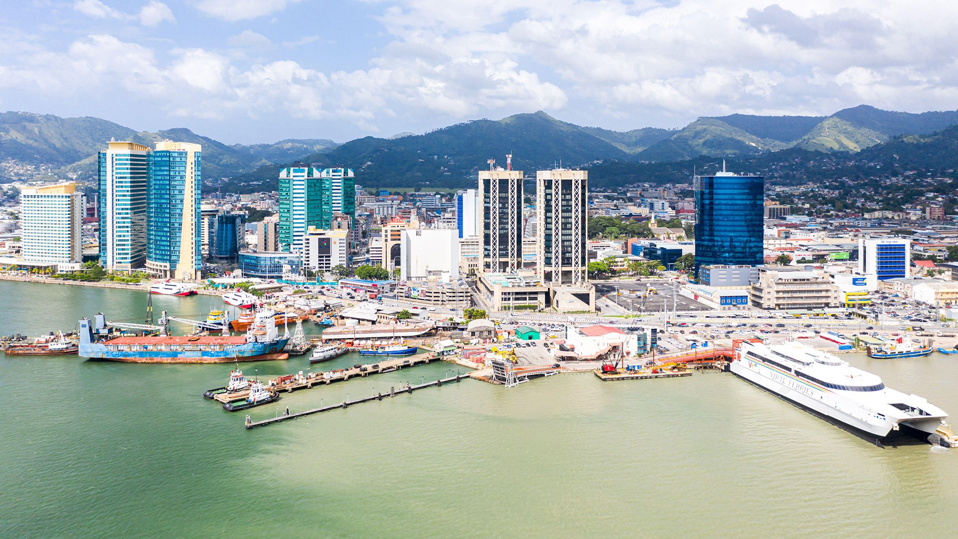 Aerial view of Port of Spain, Trinidad and Tobago, showcasing modern high-rise buildings, a bustling port with various ships docked, and lush green mountains in the background under a partly cloudy sky.