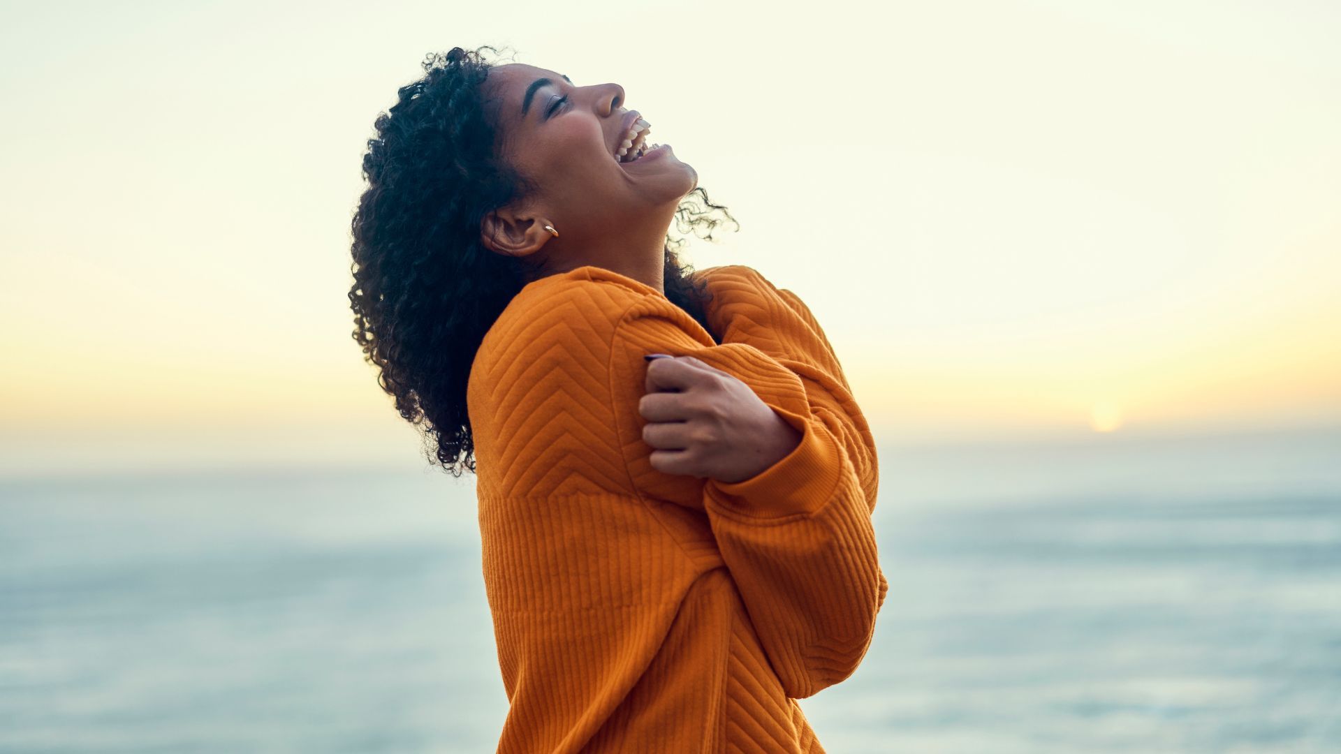 Woman in an orange sweater smiling by the ocean at sunset.